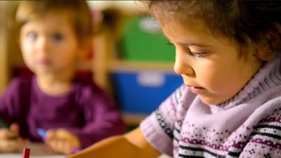 Close-up of a young girl drawing in a classroom with another child and colorful storage bins in the blurred background.