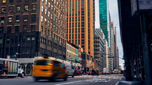 A motion-blurred yellow taxi cab drives down a busy New York City street lined with tall skyscrapers and office buildings.