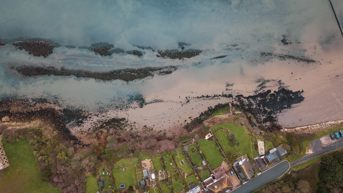 Aerial drone photograph of a coastal shoreline with houses, green lawns, a sandy beach, and shallow ocean water