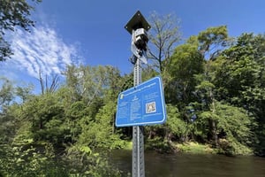 A flood sensor and blue informational sign with a QR code mounted on a metal post beside a wooded riverbank.