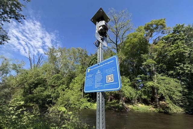 A flood sensor and blue informational sign with a QR code mounted on a metal post beside a wooded riverbank.