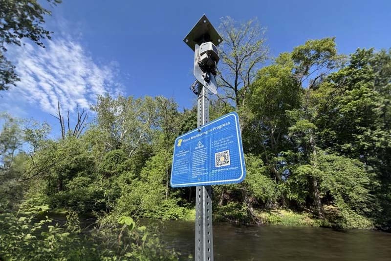 A flood sensor and blue informational sign with a QR code mounted on a metal post beside a wooded riverbank.