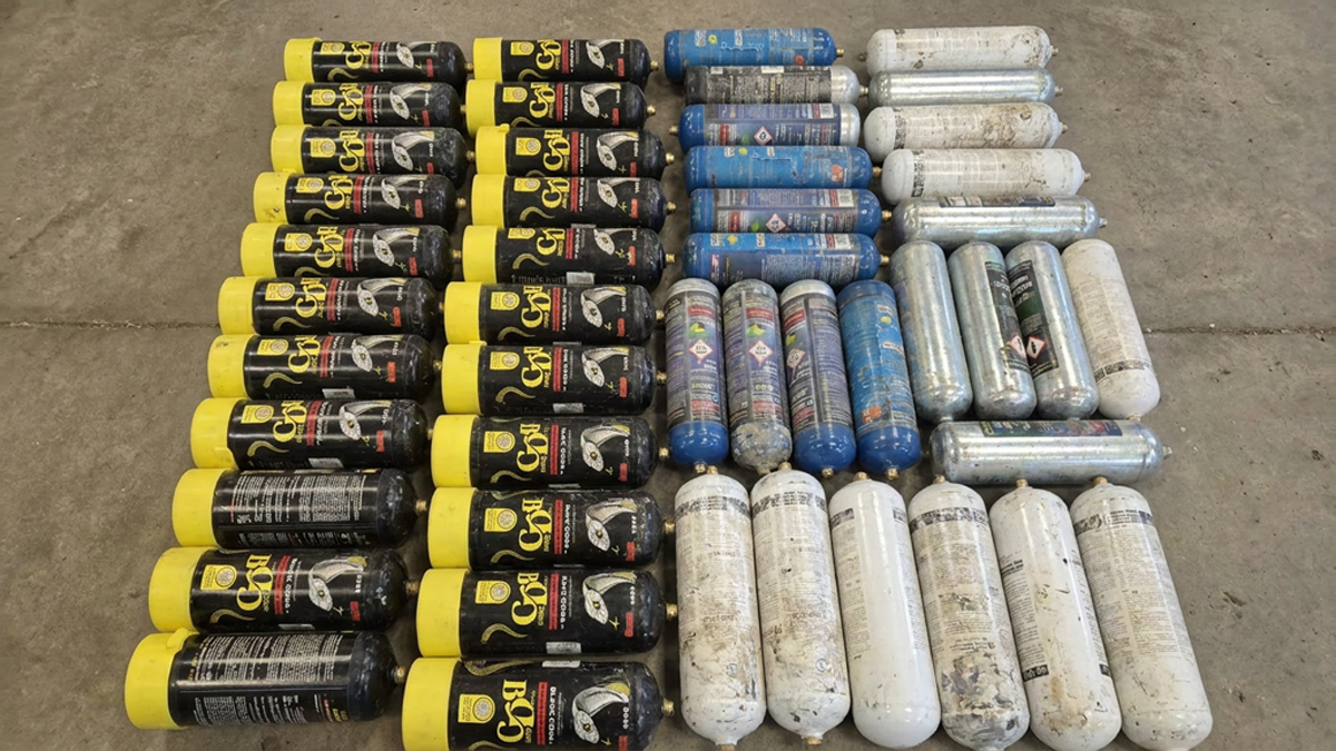 Dozens of discarded nitrous oxide canisters in black, blue, and white arranged in rows on a concrete floor