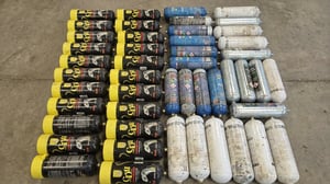 Dozens of discarded nitrous oxide canisters in black, blue, and white arranged in rows on a concrete floor