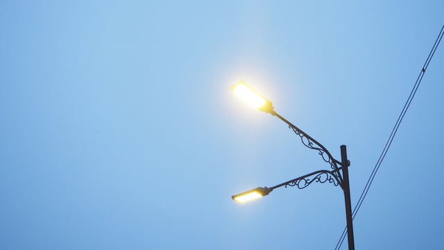 Two illuminated LED streetlights on an ornamental metal pole against a clear blue sky