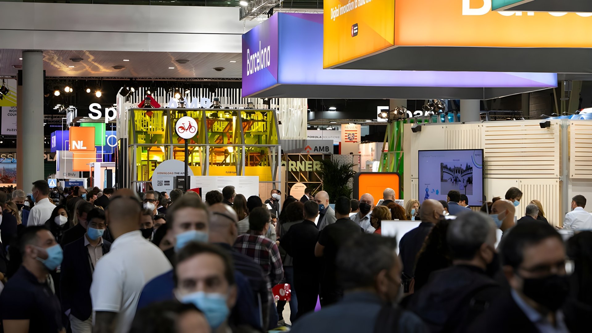 Crowded technology trade show floor with attendees in face masks walking past illuminated overhead booth signage.