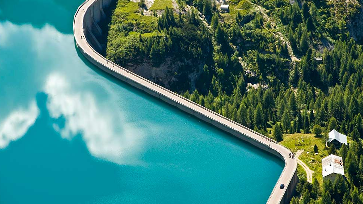 Aerial drone view of a curved concrete water dam wall bordering a turquoise reservoir and a forested mountain slope