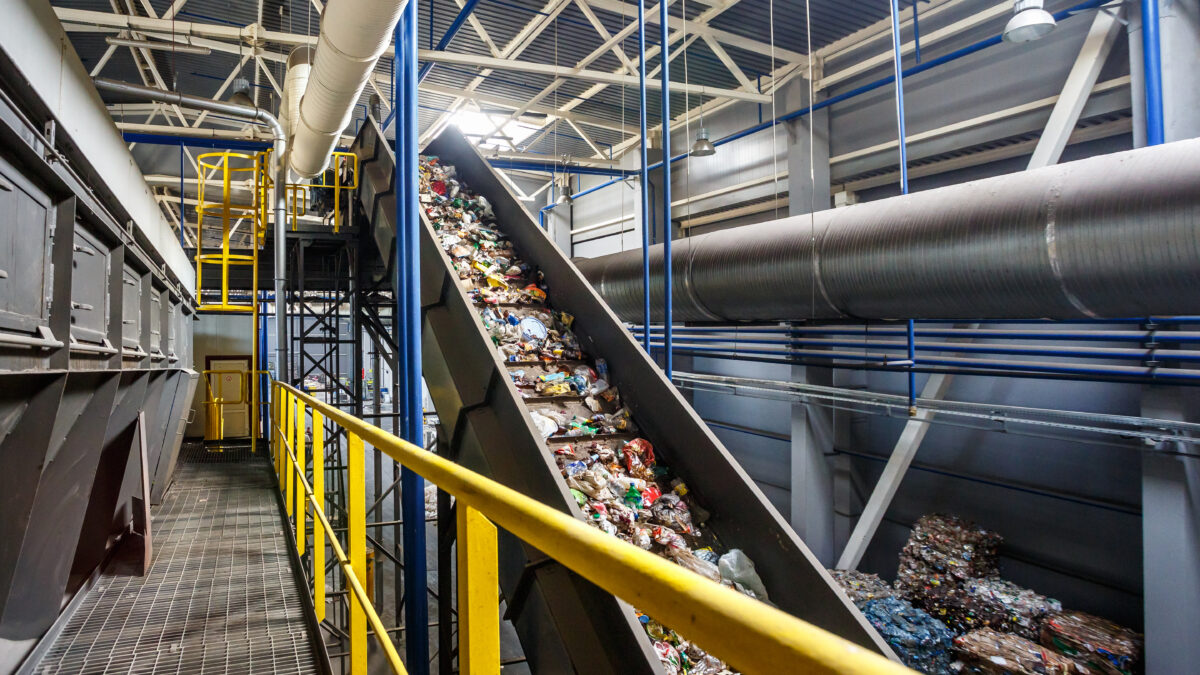 A conveyor belt in a large industrial facility carries a stream of mixed waste towards a pile of baled recyclables.