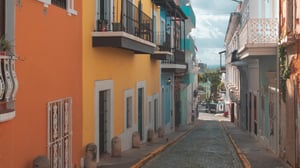 A narrow cobblestone street lined with brightly colored buildings featuring balconies and ornate railings under a partly cloudy sky.