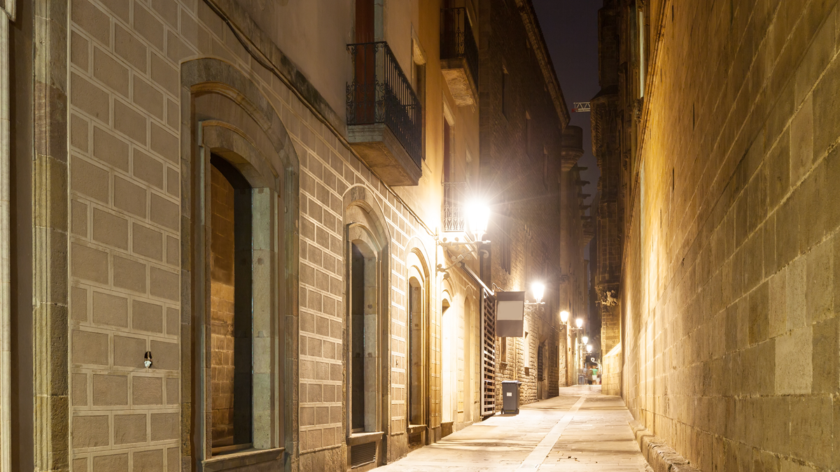 Wall-mounted streetlights illuminate a narrow stone alleyway between historic buildings at night.