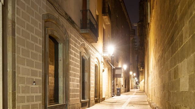 Wall-mounted streetlights illuminate a narrow stone alleyway between historic buildings at night.