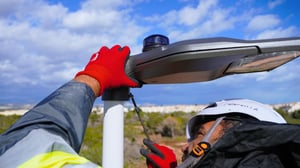 A technician wearing red gloves and a hard hat installs an NB-IoT node onto a modern street light fixture outdoors