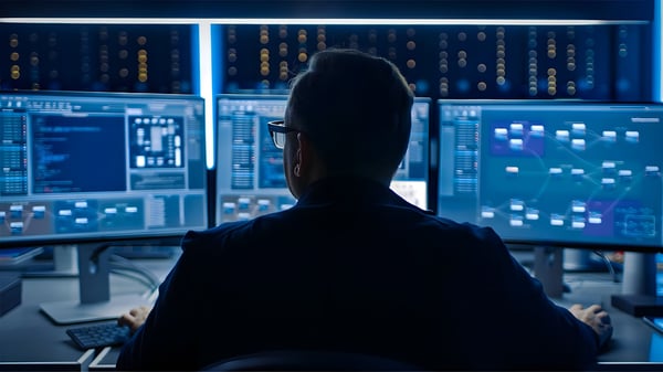 Rear view of an IT professional monitoring network diagrams and data code on multiple computer screens in a dark room.