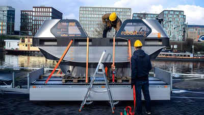 Two workers in hard hats secure a Roboat.org autonomous vessel to a trailer on a waterfront with city buildings behind.