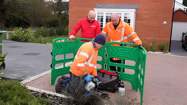 Three Severn Trent workers in high-visibility jackets install a water meter behind a green safety barrier on a driveway.