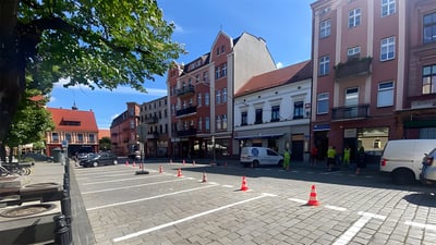 Cobblestone parking lot in Ostrow Wielkopolski with traffic cones, white vans, and workers in high-visibility vests.
