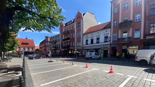 Cobblestone parking lot in Ostrow Wielkopolski with traffic cones, white vans, and workers in high-visibility vests.