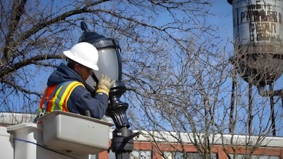 A technician in a bucket truck installs a decorative LED street lamp near a water tower and bare trees.