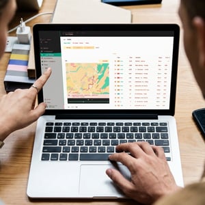 Two people viewing a laptop screen displaying a digital map and data dashboard on a wooden desk