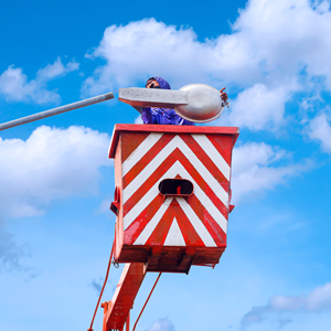 A technician in a cherry picker bucket installs a street light fixture against a blue sky with clouds.