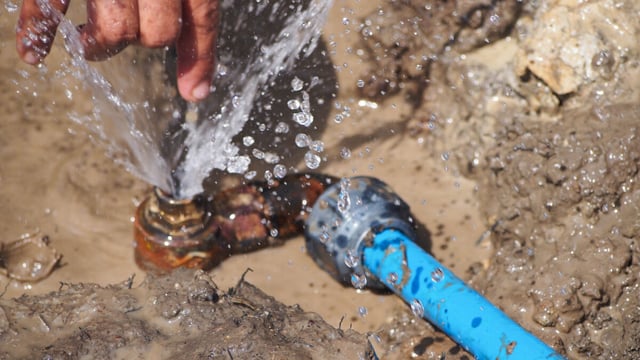 Water spraying from a leaking corroded pipe joint connected to a blue pipe, with a hand above and wet mud around it.