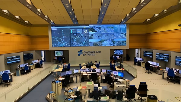 Operators at desks in a Dirección General de Tráfico control room with large wall monitors displaying road traffic data.