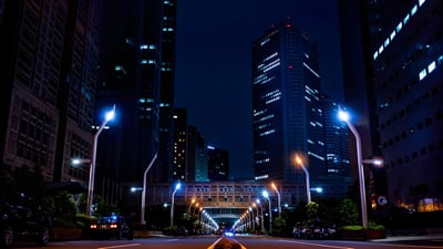 Modern LED street lights illuminate a city road at night between dark skyscrapers.