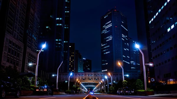 Modern LED street lights illuminate a city road at night between dark skyscrapers.