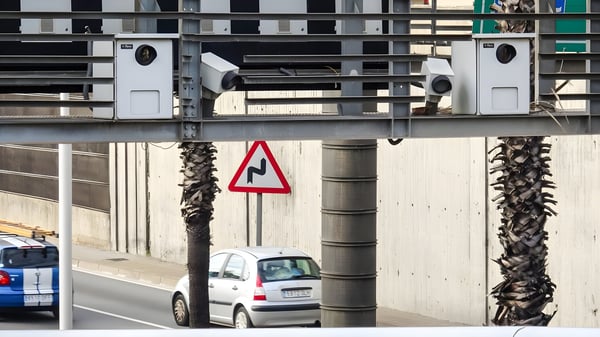 Four traffic surveillance cameras mounted on a metal gantry over a highway with passing cars and a winding road sign.