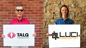 Two people holding signs with the TALQ Consortium and LUCI Association logos in front of brick and stone walls