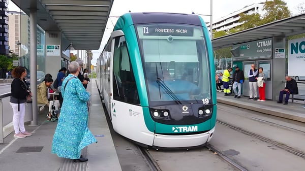 A green and white TRAM Barcelona light rail vehicle arrives at a covered station platform with waiting passengers.