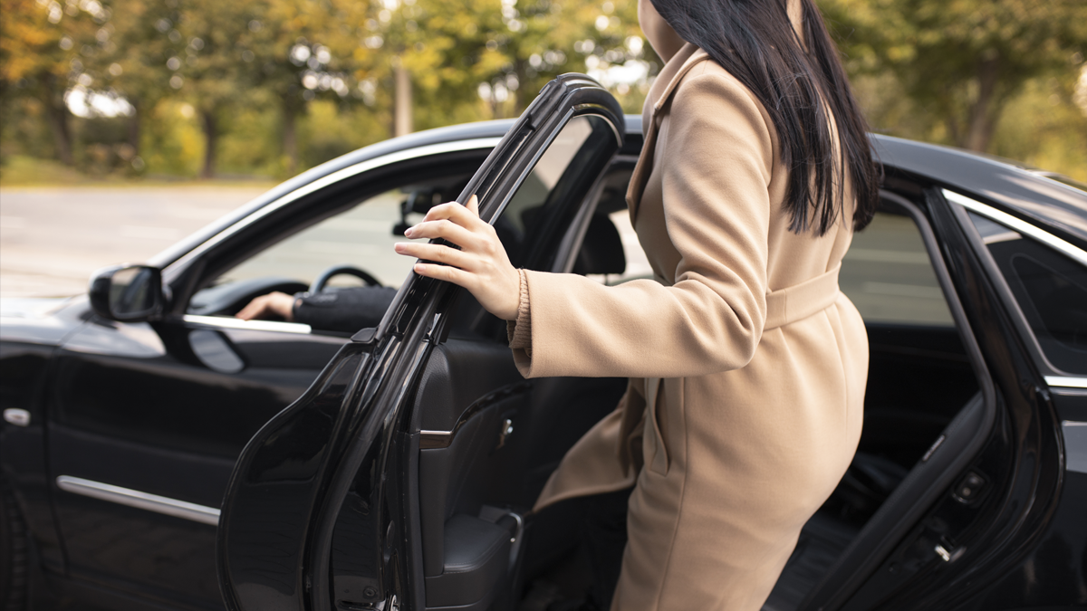 Woman in a beige coat entering the open rear door of a black sedan outdoors.