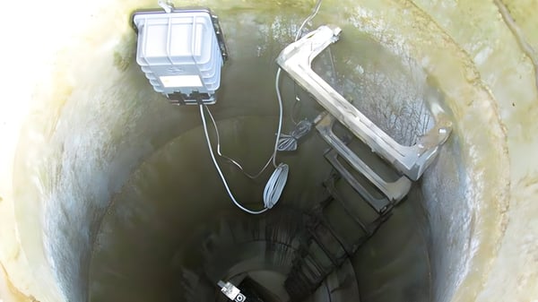 A grey IoT sensor device and wiring mounted inside a concrete manhole shaft with a metal access ladder.