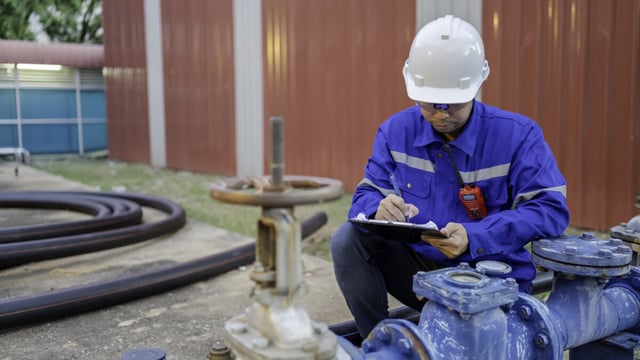 Engineer in a white hard hat and blue overalls writing on a clipboard beside blue water pipework, valves and a handwheel
