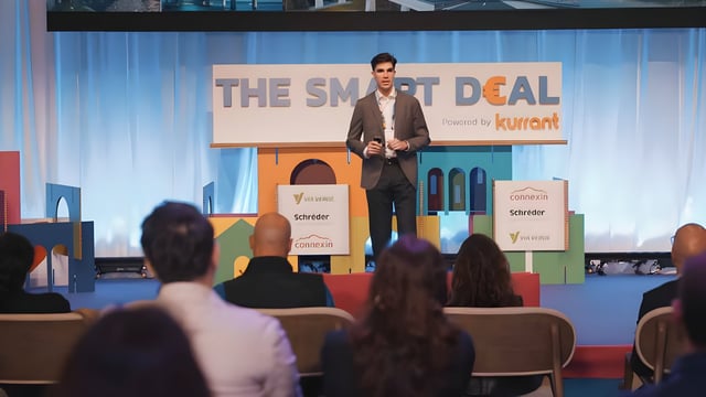 A man presents on a stage with "The Smart Deal powered by Kurrant" signage before an audience at a conference.