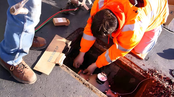 A utility worker in a high-visibility orange jacket installs a smart water meter inside an underground street vault.