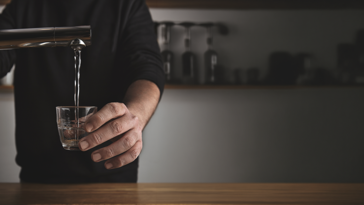 A person filling a small glass with water from a modern kitchen tap over a wooden countertop