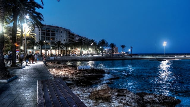 Illuminated coastal promenade in Torrevieja at night with streetlights, palm trees, pedestrians, and ocean water