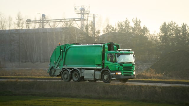 Green rear-loading garbage truck driving along a rural road at dawn with industrial structures and trees in the background
