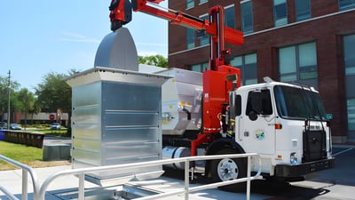 A white truck with a red hydraulic crane lifts a large metal underground waste container outside a brick building.