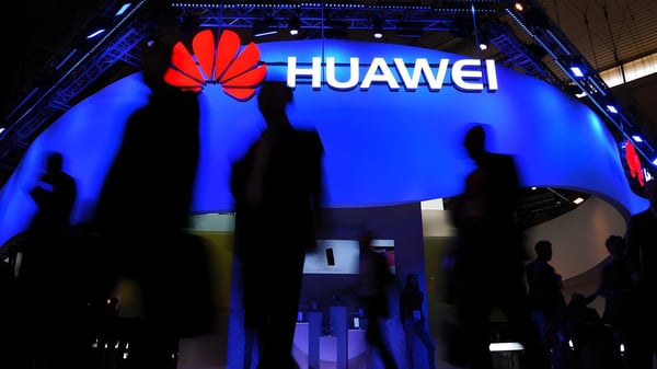 Silhouetted people walk past a glowing blue Huawei exhibition booth featuring the company's red flower logo.