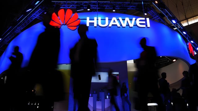 Silhouetted people walk past a glowing blue Huawei exhibition booth featuring the company's red flower logo.