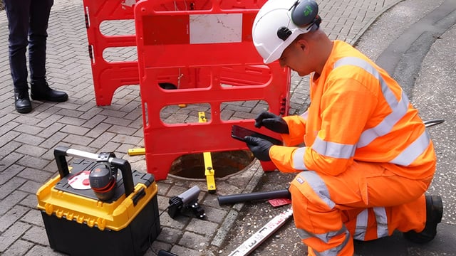 A utility worker in high-visibility gear uses a tablet near an open manhole with red safety barriers and monitoring tools.
