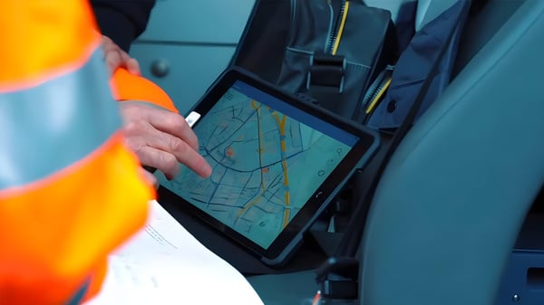 A utility worker in high-visibility clothing uses a tablet displaying a digital city map inside a vehicle.