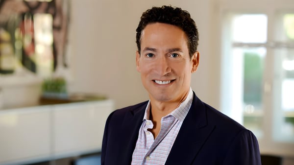 A man with dark curly hair wearing a navy blazer and patterned button-down shirt in a bright indoor office setting.