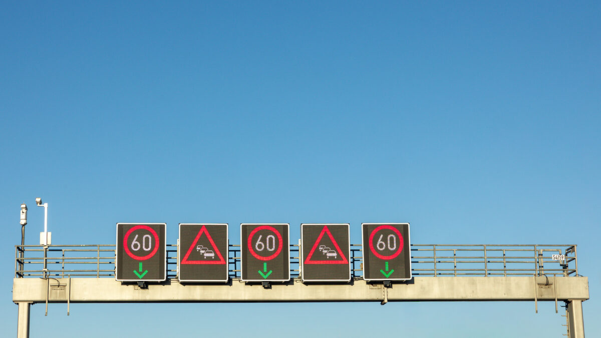 Overhead gantry with five digital road signs displaying alternating 60 km/h speed limits and vehicle incident warnings under a clear blue sky.