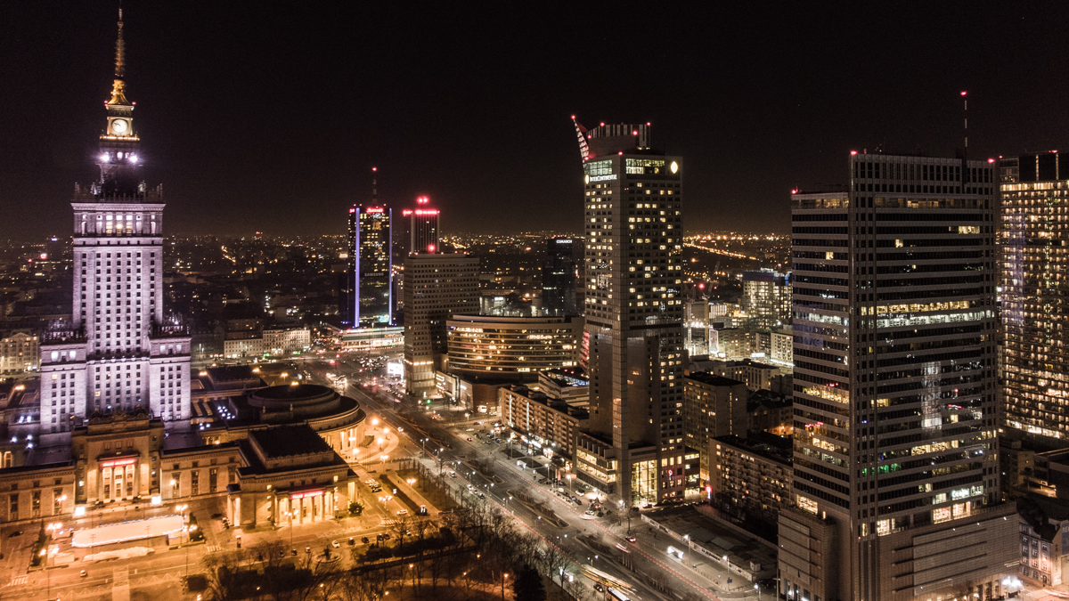 Nighttime skyline of illuminated Warsaw featuring the Palace of Culture and Science alongside modern skyscrapers.