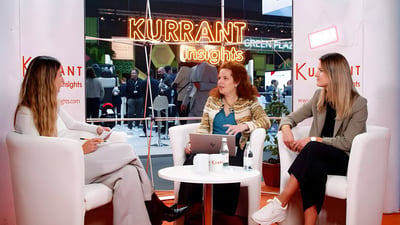 Three women sit in white armchairs for a Kurrant Insights interview at a trade show with a neon sign in the background.