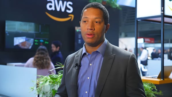 A man in a suit jacket speaks in front of an AWS branded booth at a technology trade show.