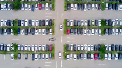 Top-down aerial view of a large outdoor parking lot with rows of cars separated by green grass medians and white arrows.